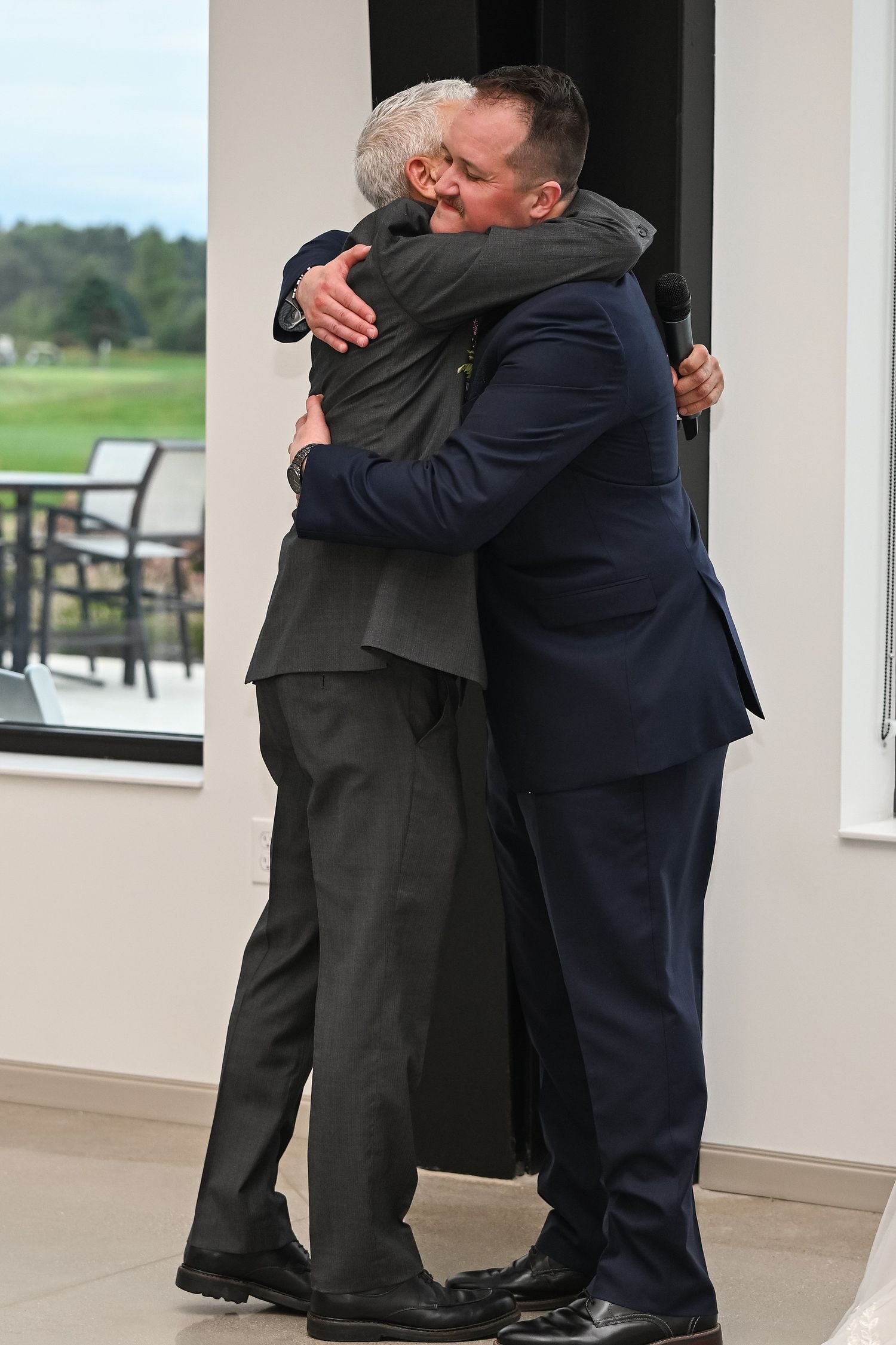 Two men share a heartfelt hug during a wedding ceremony at an elegant venue with golf course views.