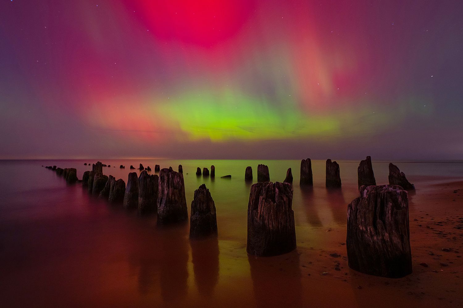 Vibrant aurora borealis lights dance across the night sky over old wooden pier posts in calm water.
