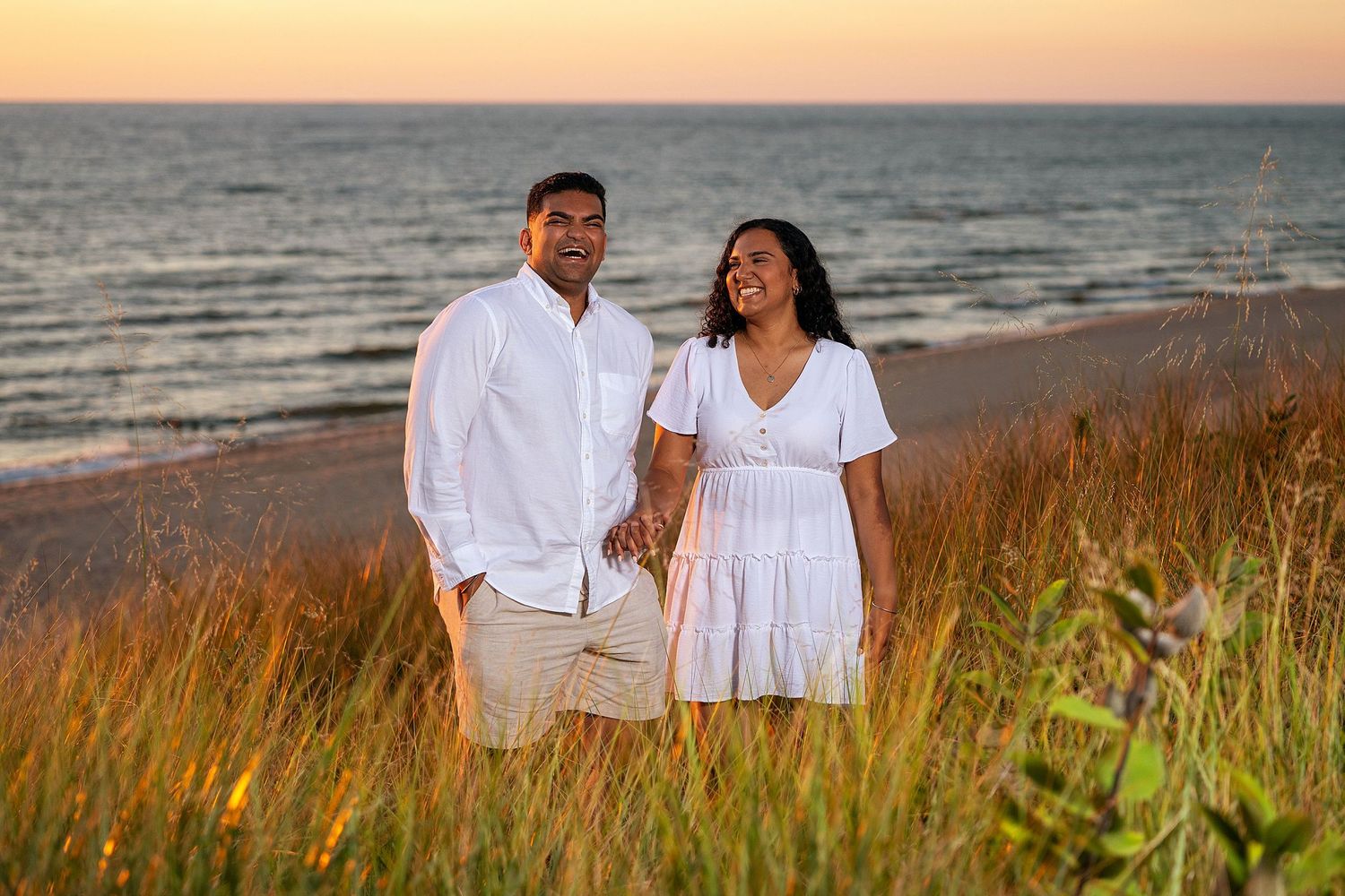 Couple walking along beach at sunset through golden grass.