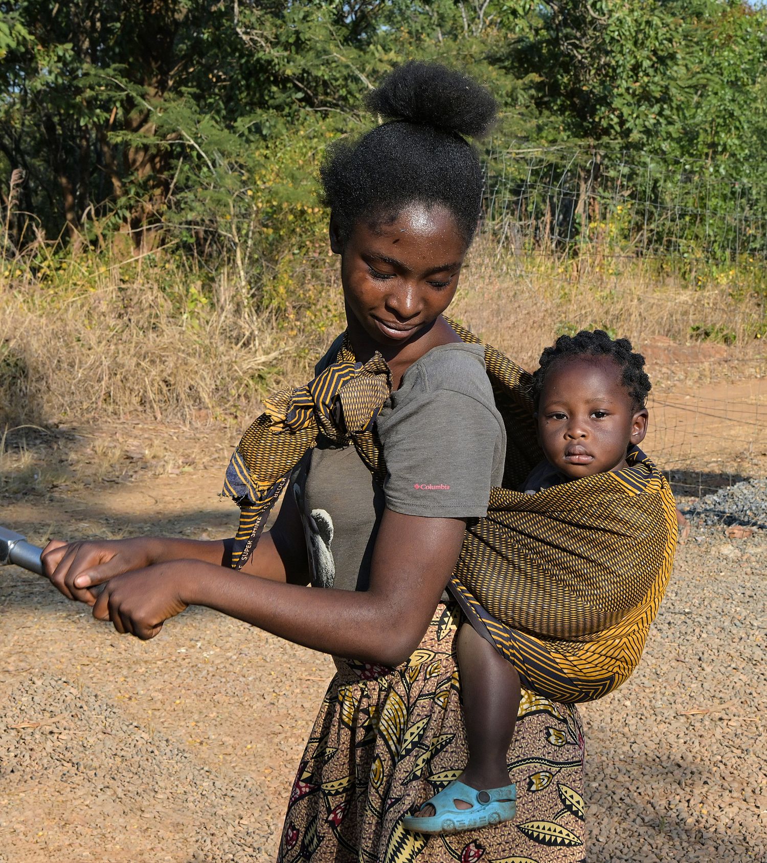 A woman carries a small child in a traditional wrap sling on a dusty path.