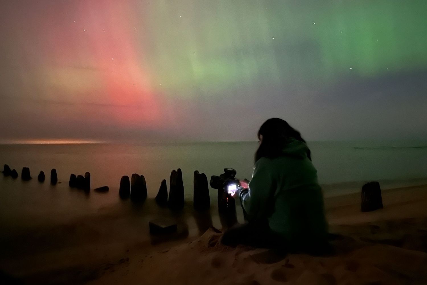 Woman sitting on a sandy beach photographing vibrant aurora borealis lights in green and red hues over calm water at night.