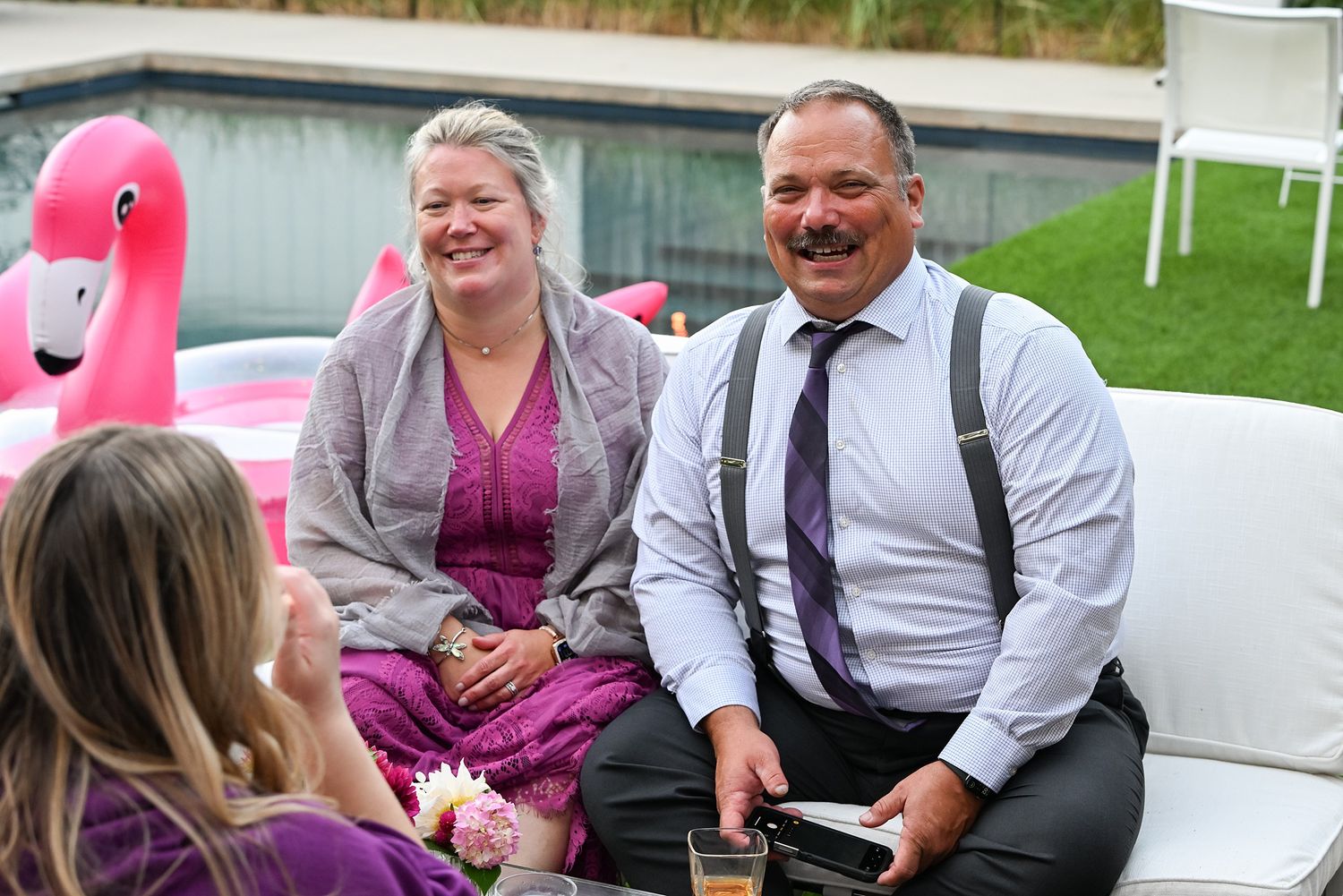Wedding guests share a laugh while sitting outdoors with a pink flamingo decoration visible in the background.