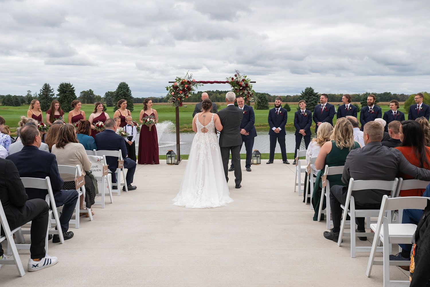 Bride walking down outdoor aisle toward groom with wedding party standing at floral arch backdrop.
