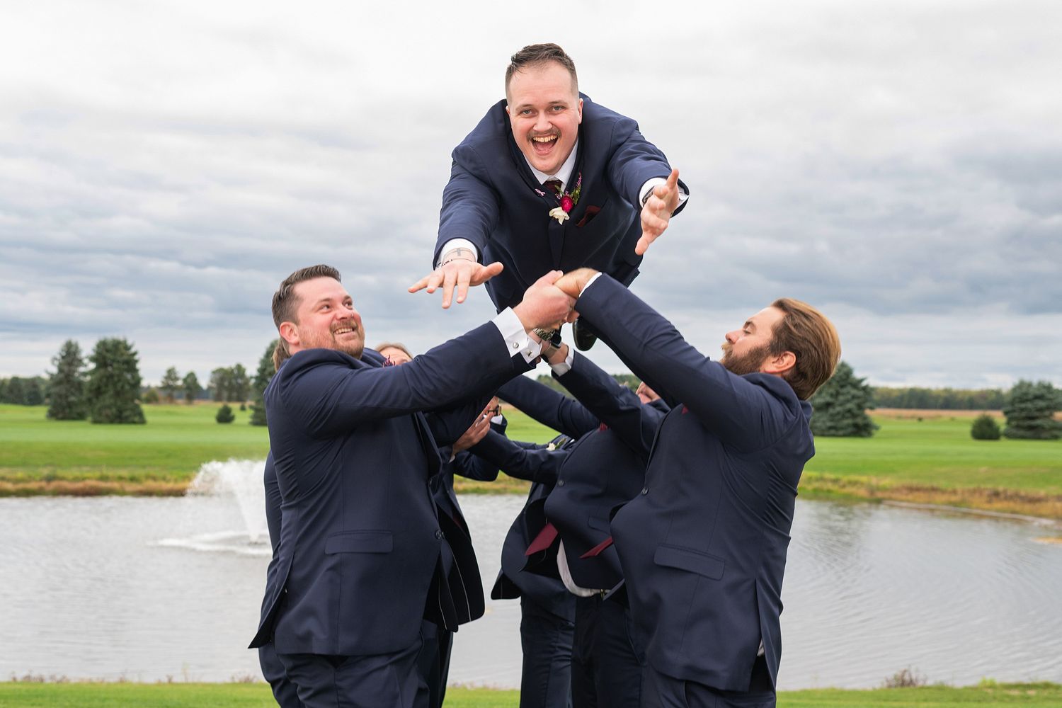 Groomsmen tossing groom in the air outdoors at a golf course wedding venue.