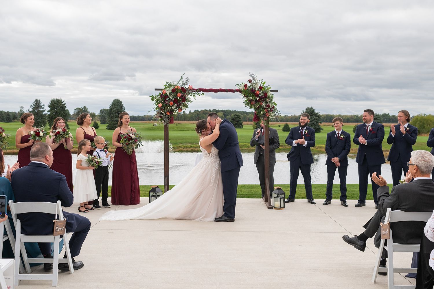 Bride and groom share first kiss under floral arch at outdoor waterfront wedding ceremony.