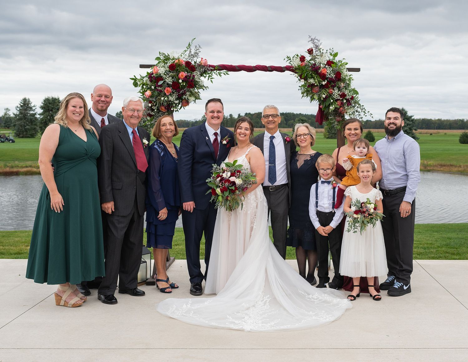 Wedding party and family pose together outdoors at ceremony arch with floral decorations near pond.