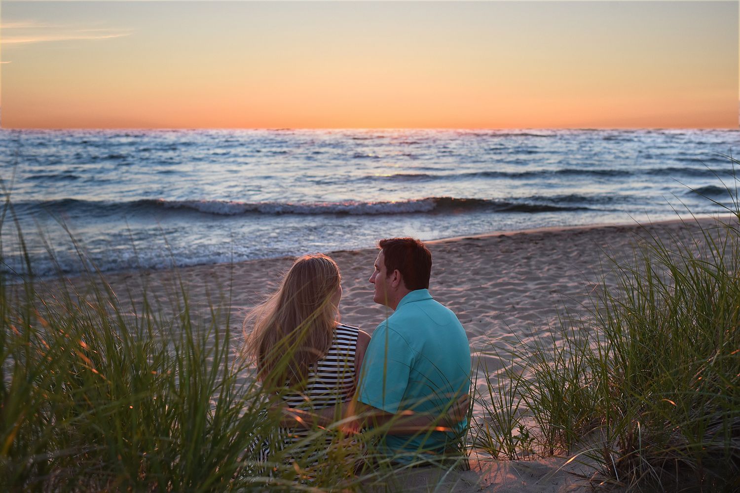 Couple sitting on sandy beach dunes watching a colorful sunset over calm ocean waves at dusk.