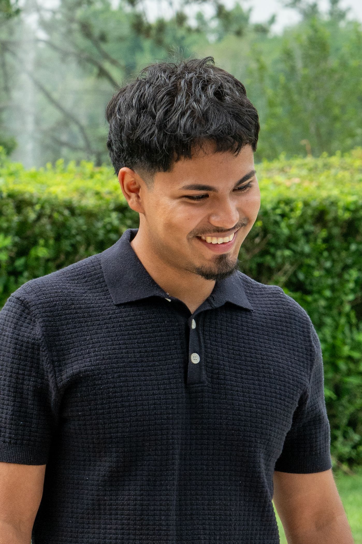 Smiling young man with dark curly hair wearing a black polo shirt standing outdoors near green hedges.