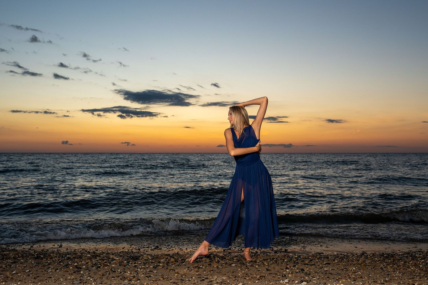 Silhouetted dancer strikes artistic pose on beach during dramatic sunset over water.