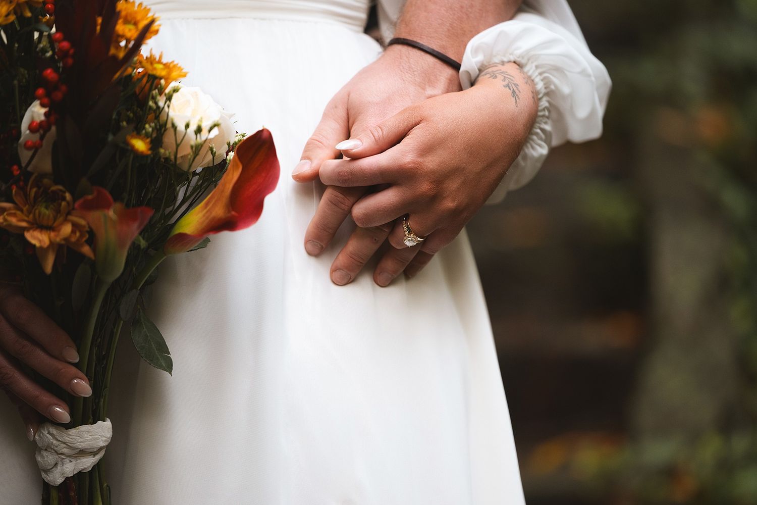 Close-up of a bride and groom holding hands with wedding rings, beside an autumn bouquet of orange and burgundy flowers.