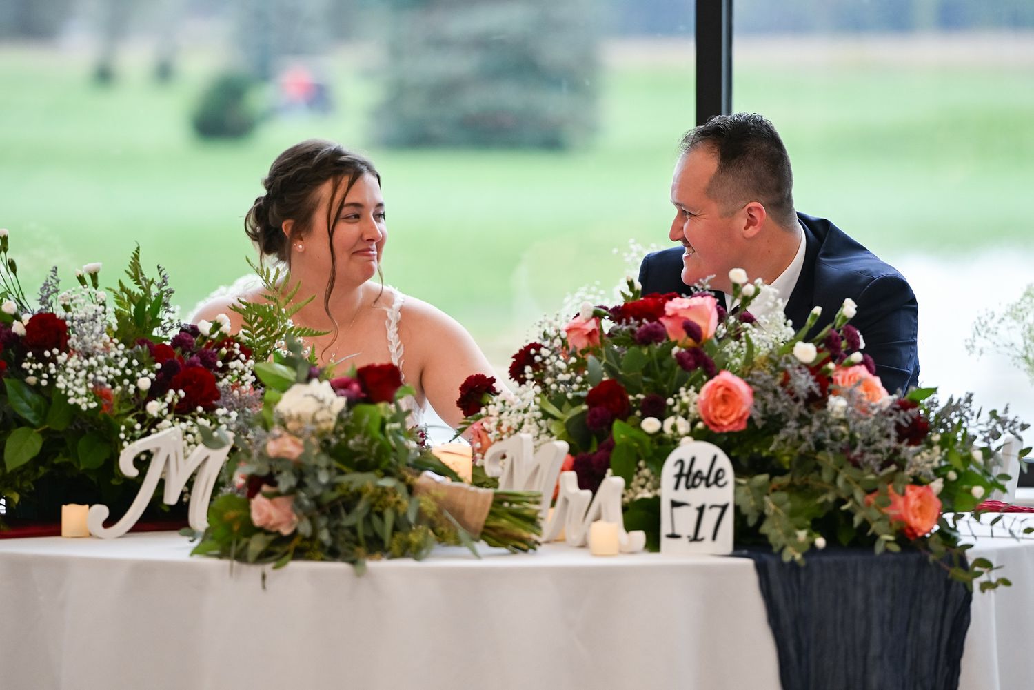 Bride and groom smile at sweetheart table adorned with red floral arrangements.