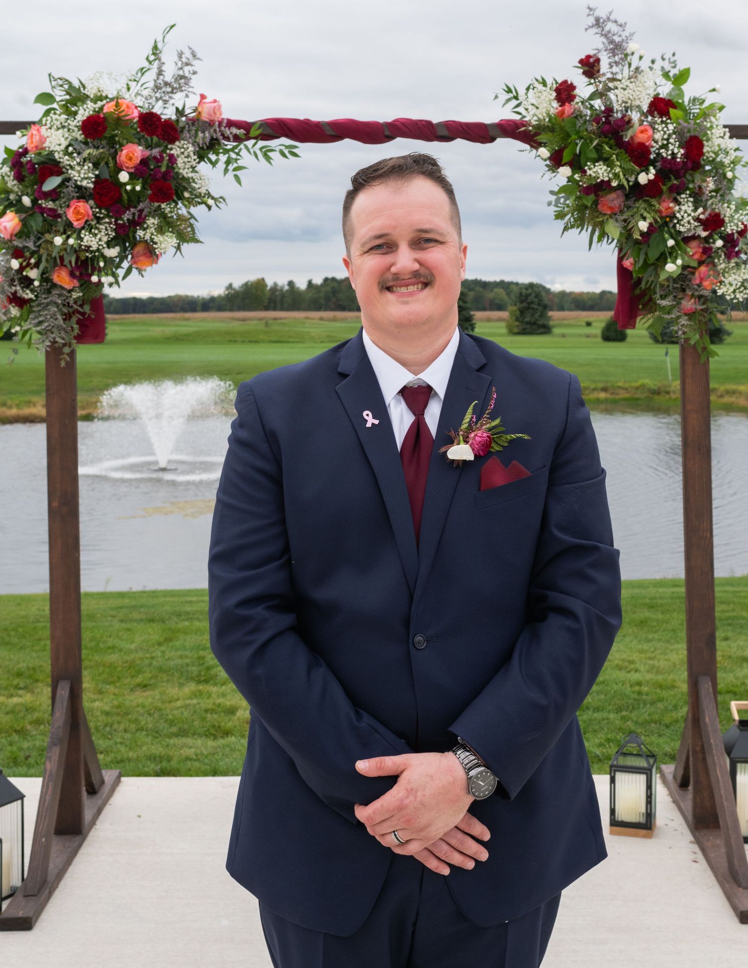Groom in navy suit with burgundy tie and boutonniere stands under floral arch at outdoor wedding venue.