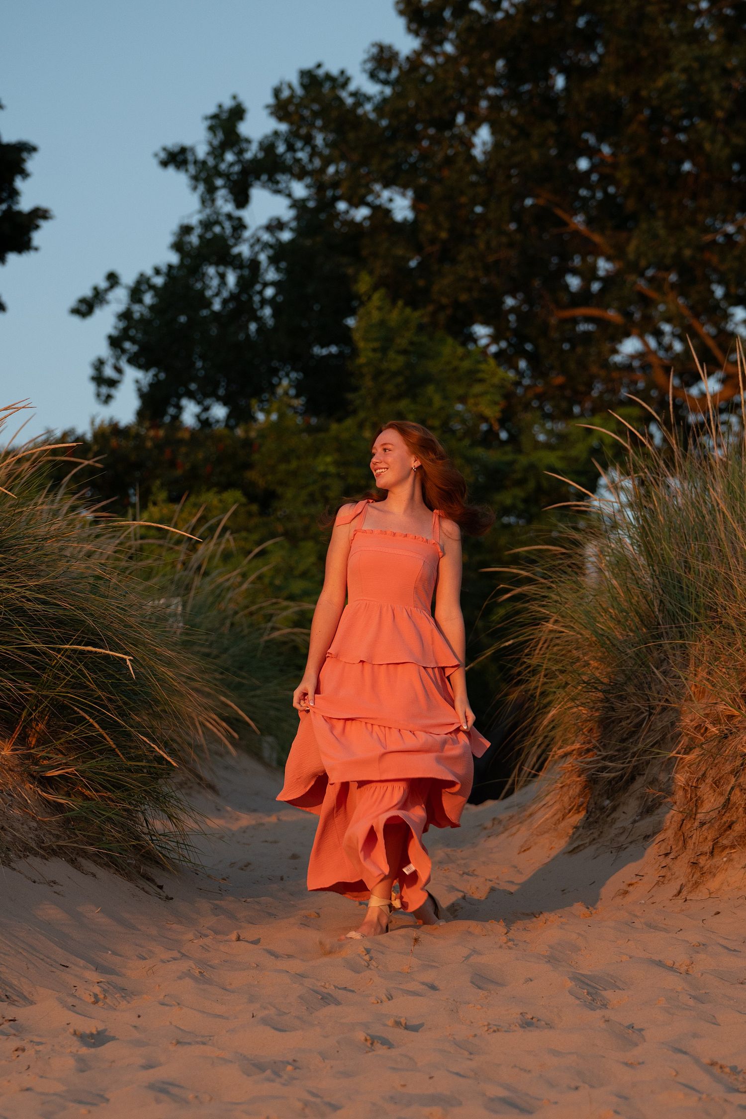 Woman in coral tiered dress walking through sandy dunes at golden hour, with green trees in background.