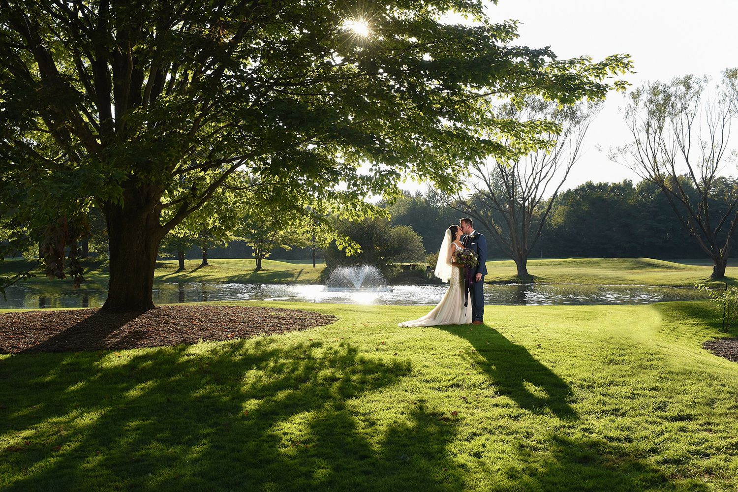Wedding couple silhouetted against a sunset by a fountain on manicured golf course grounds.