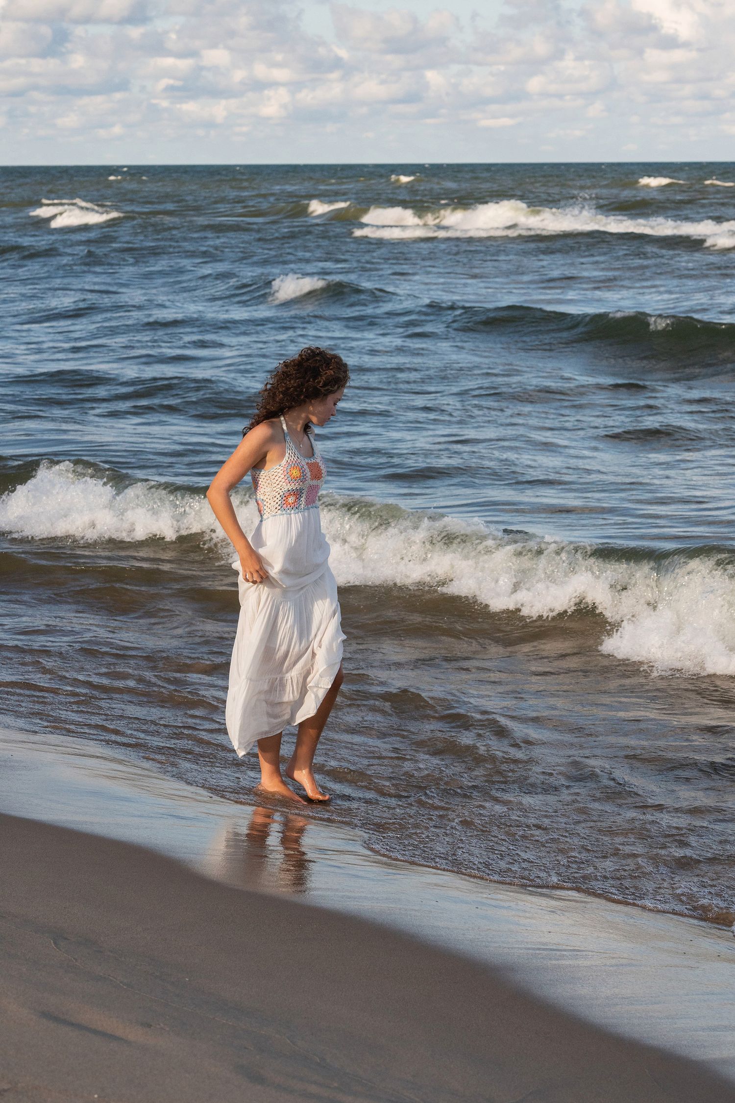 Figure in white dress explores the water's edge on a breezy beach day.