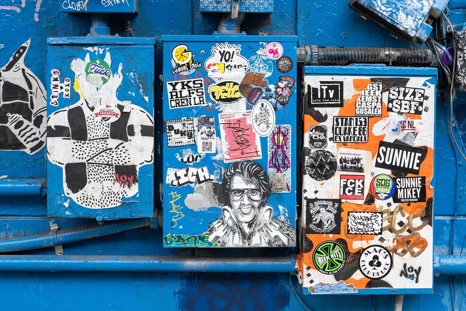 Urban wall covered in layers of graffiti stickers and street art in blue and white tones.