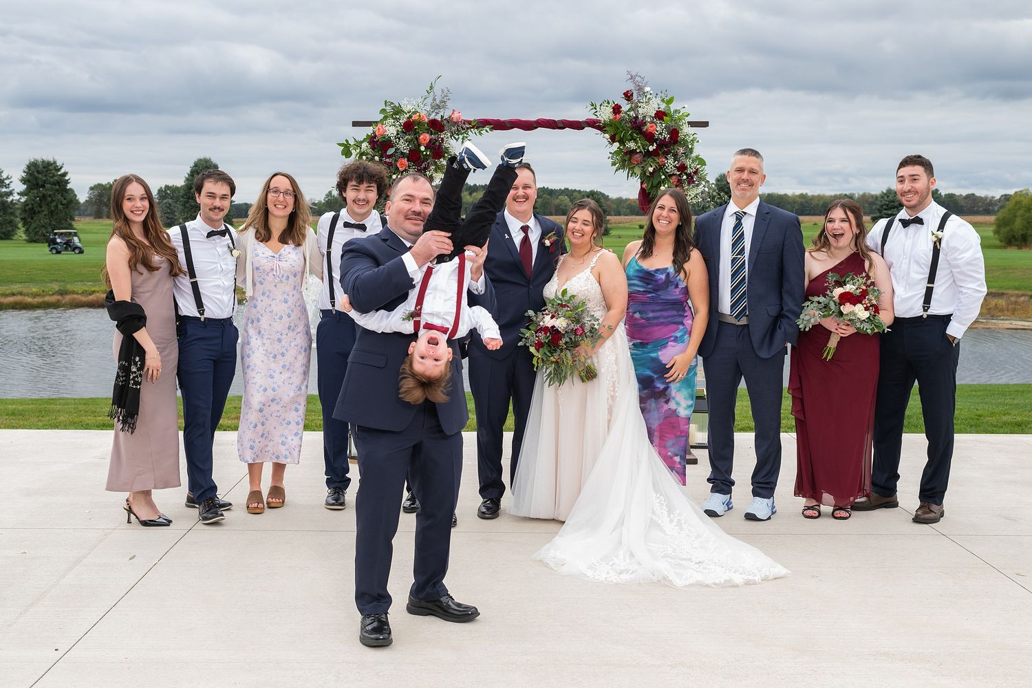 Wedding party poses outdoors near floral arch and pond, groom playfully holds child upside down while guests smile.