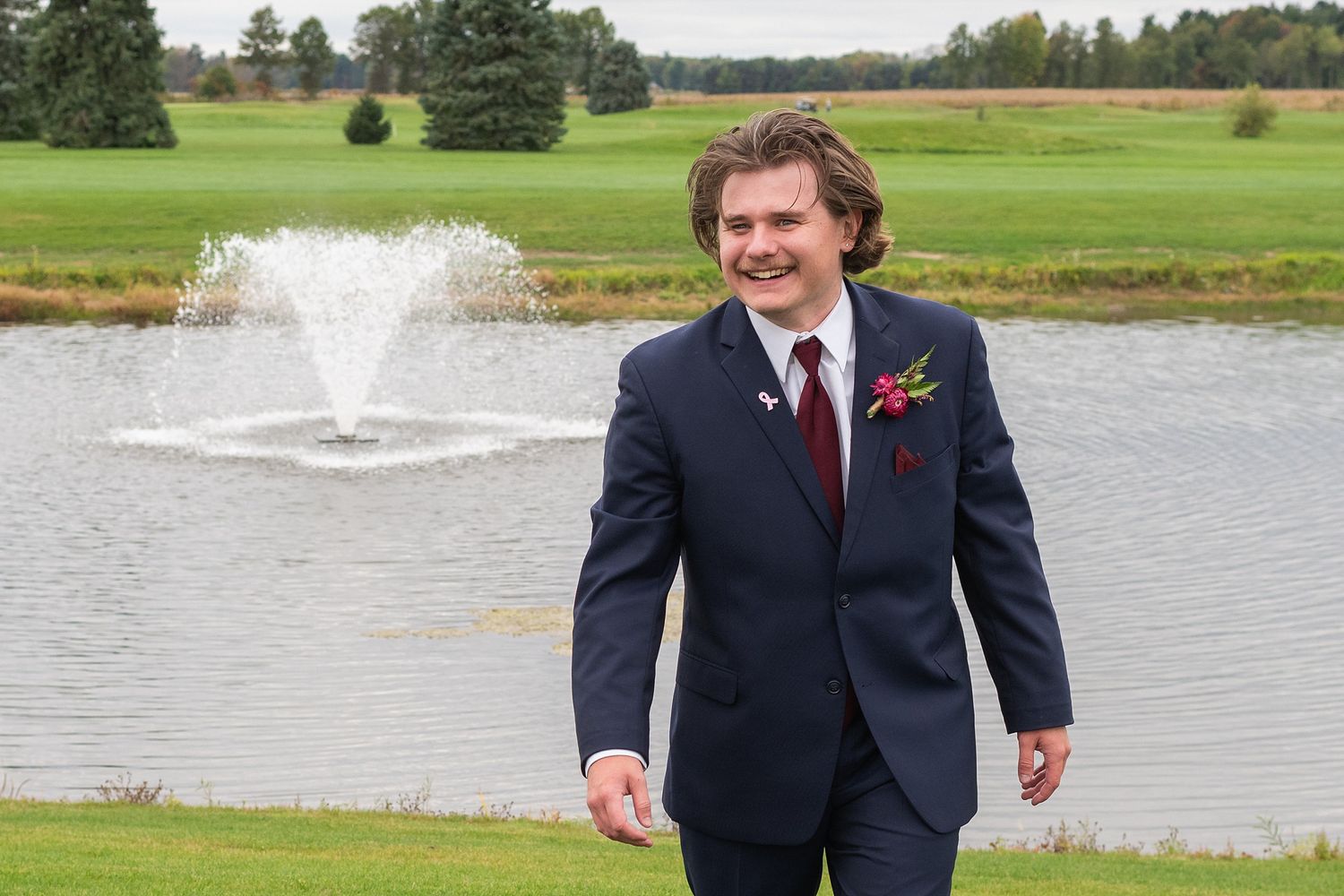 Smiling groom in navy suit with burgundy boutonniere standing by a fountain on a golf course wedding day.