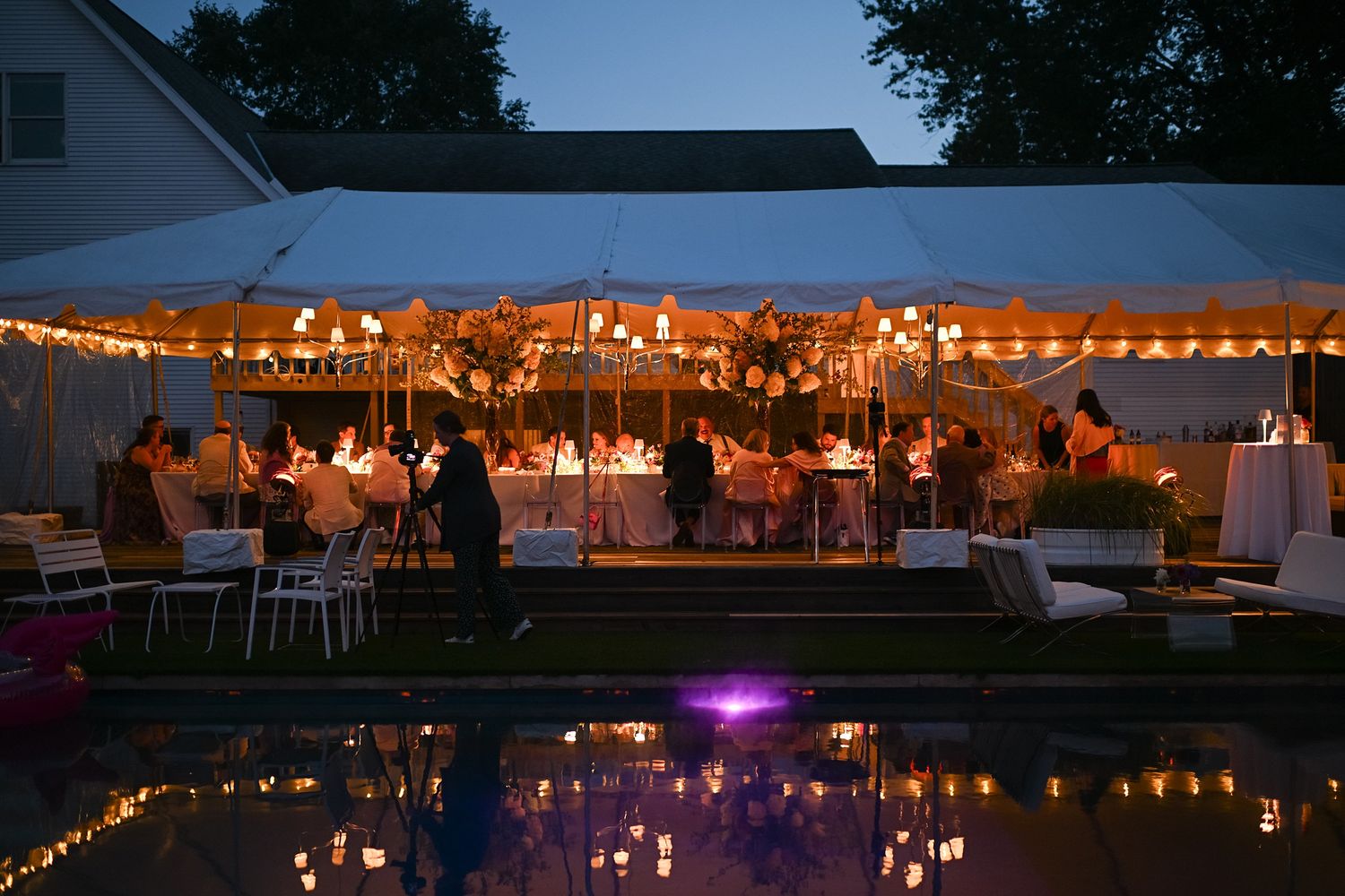 Evening outdoor wedding reception under a white tent with string lights reflecting in a pool at dusk.