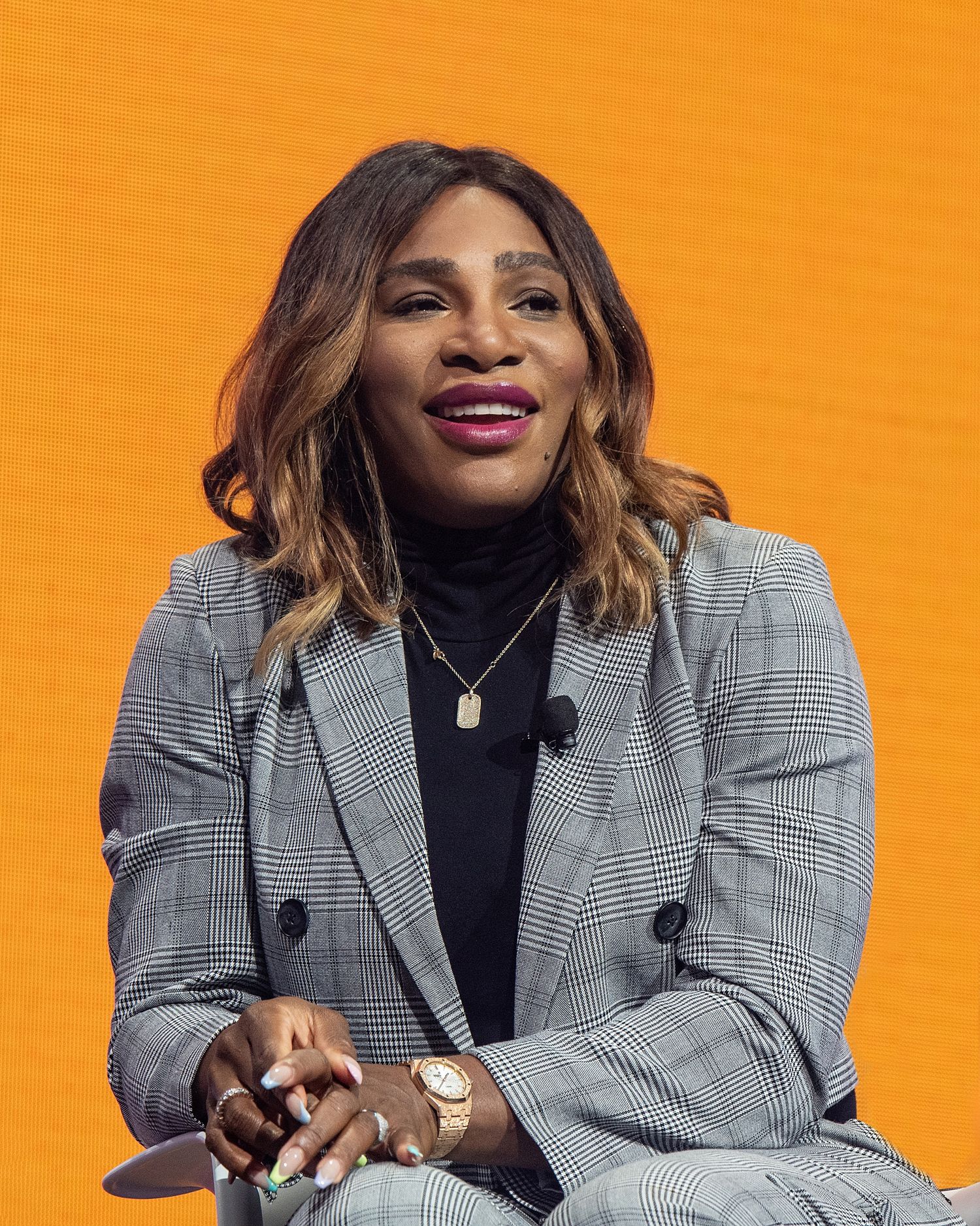 Professional speaker in plaid blazer and black turtleneck against bright orange background.