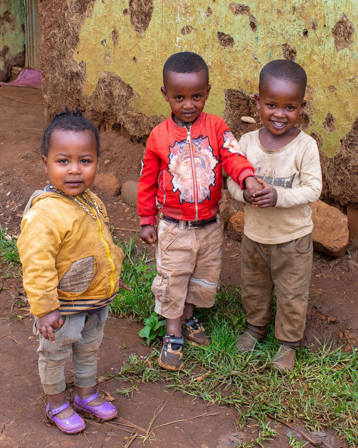 Three young children stand together against a weathered yellow wall wearing casual clothes.