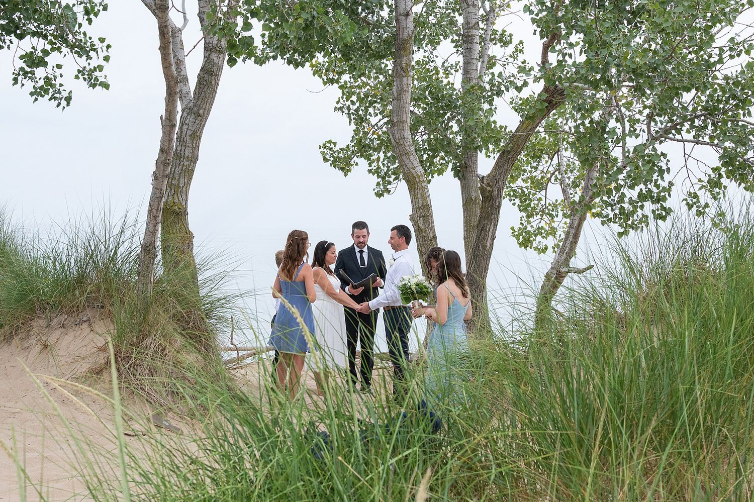 Small outdoor beach wedding ceremony among sand dunes and trees with couple and guests.
