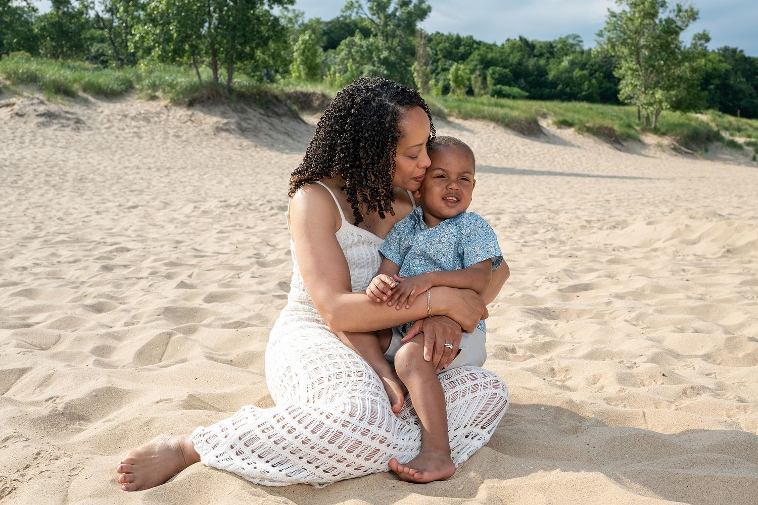A mother sits on sandy beach dunes, lovingly kissing her baby boy in a blue outfit while wearing a white lace dress.