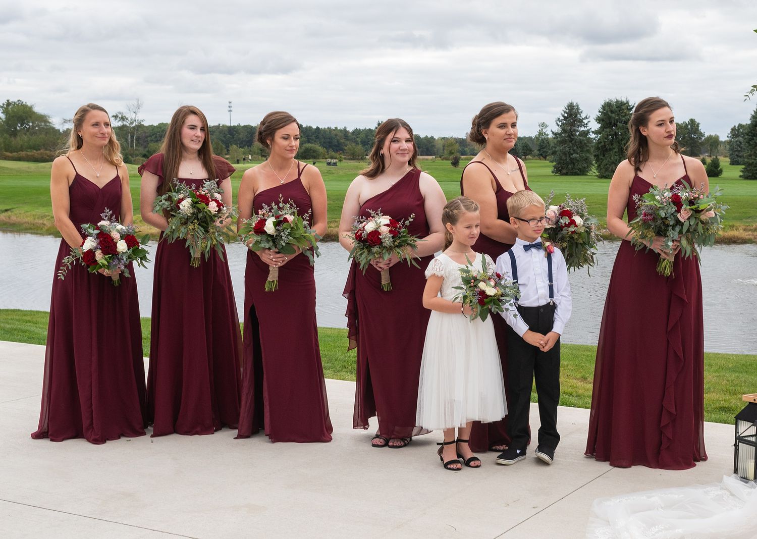 Six bridesmaids in burgundy gowns with flower girl and ring bearer at outdoor wedding.
