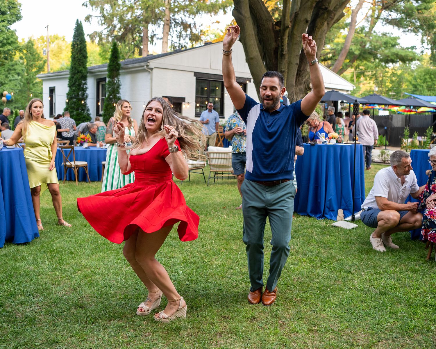 Two people dance joyfully on a lawn during an outdoor party with blue-clothed tables and guests in the background.