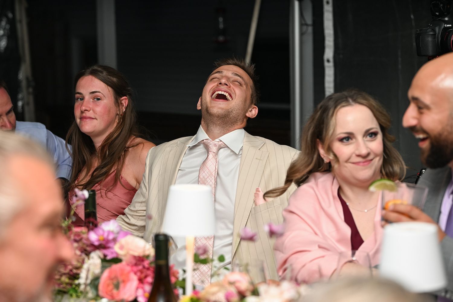 Wedding guests share a joyful moment of laughter during reception dinner with pink floral centerpieces on table.