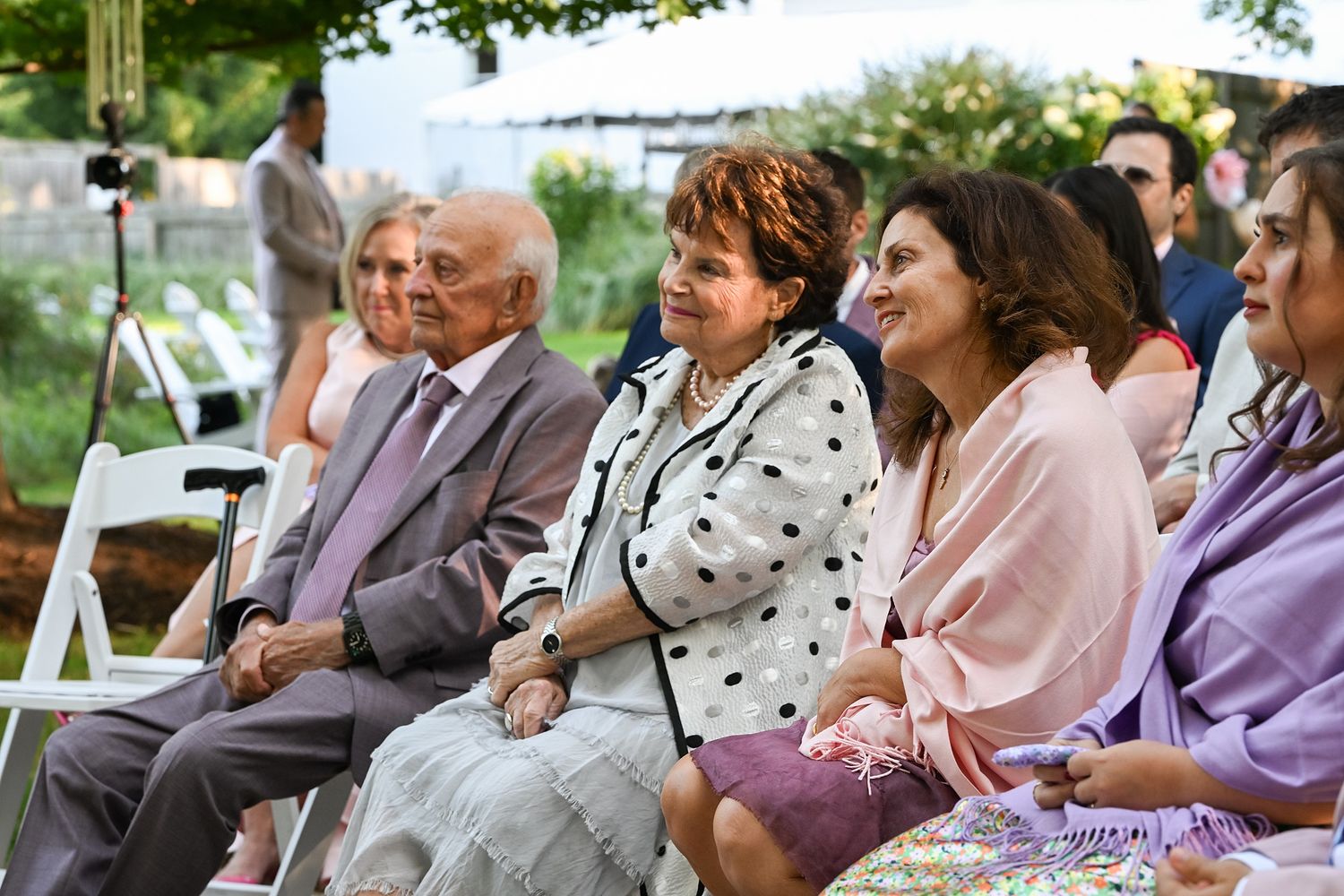 Guests seated outdoors at a wedding ceremony listen attentively to the proceedings on a sunny afternoon.