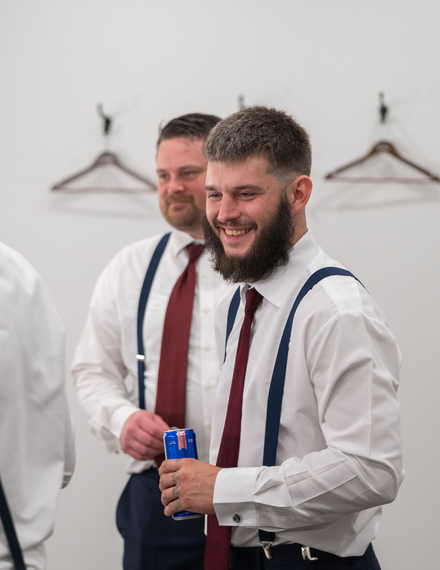 Two smiling groomsmen in white shirts, suspenders, and burgundy ties preparing for a wedding ceremony.