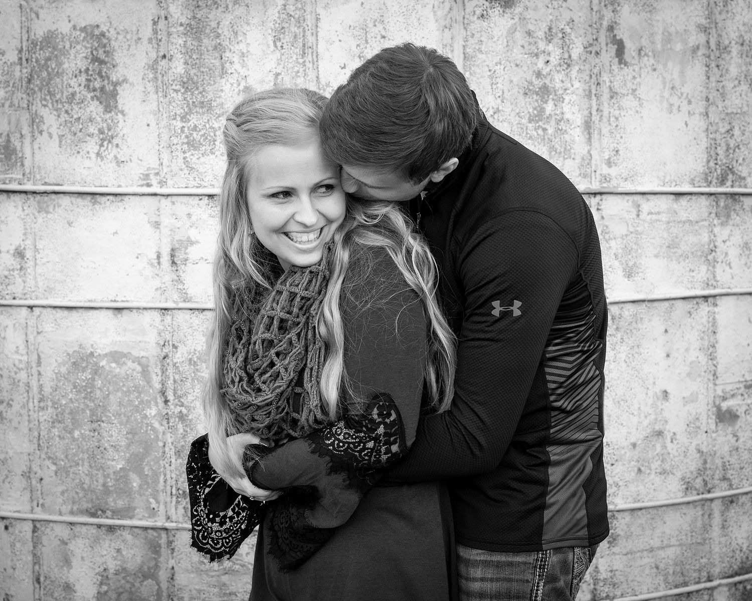 Black and white photo of a smiling couple embracing, woman laughing as man hugs her from behind outdoors.
