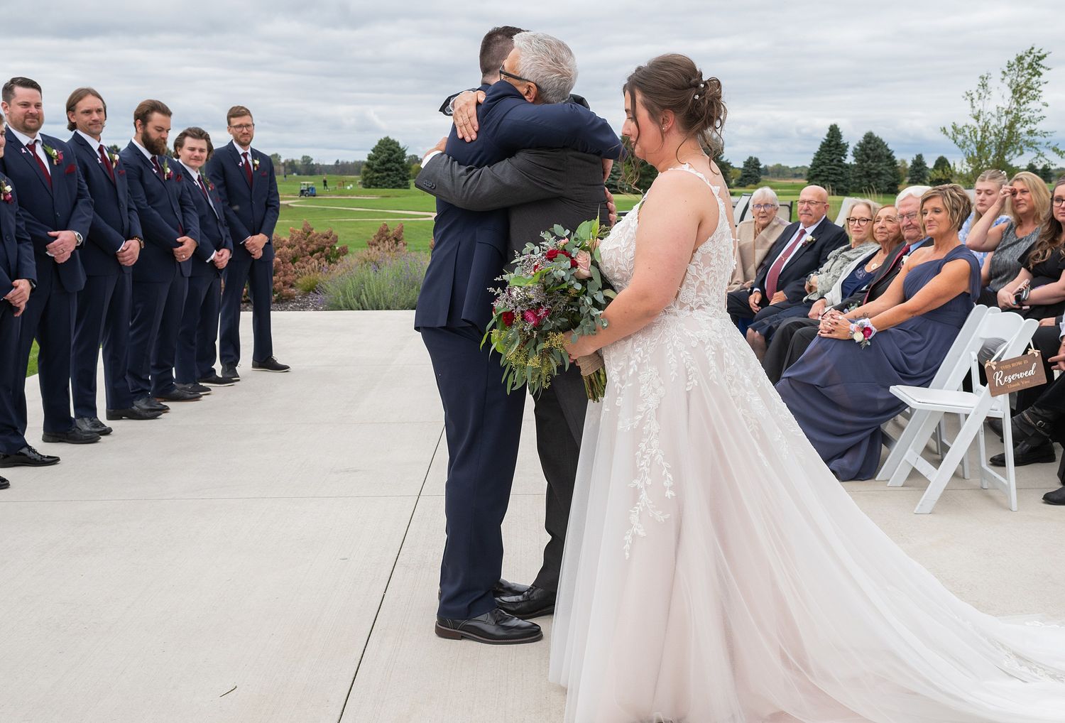 Bride in white lace gown shares emotional hug with father at outdoor wedding ceremony.