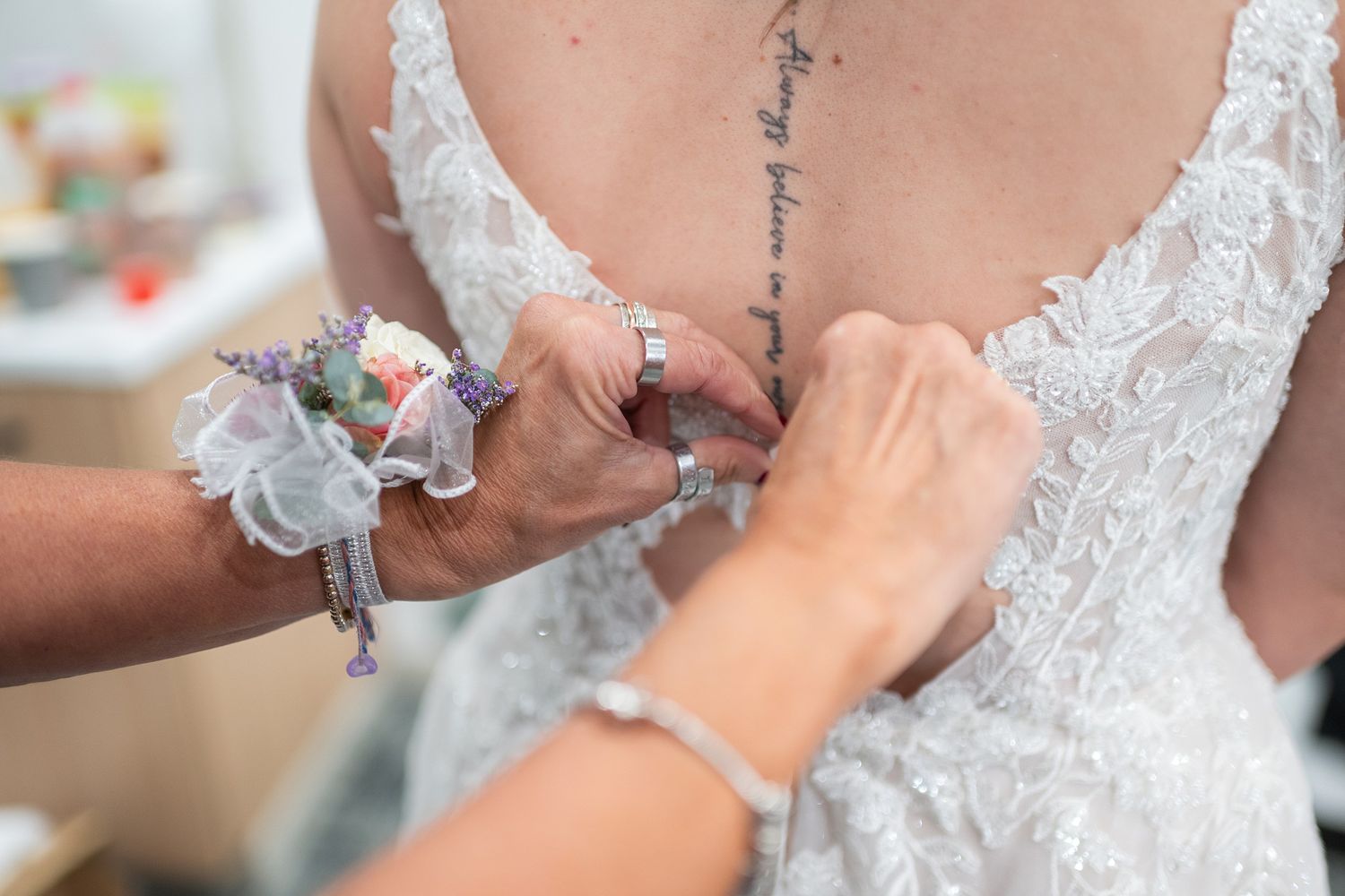 Hands fastening lace wedding dress buttons on bride with script tattoo on back.
