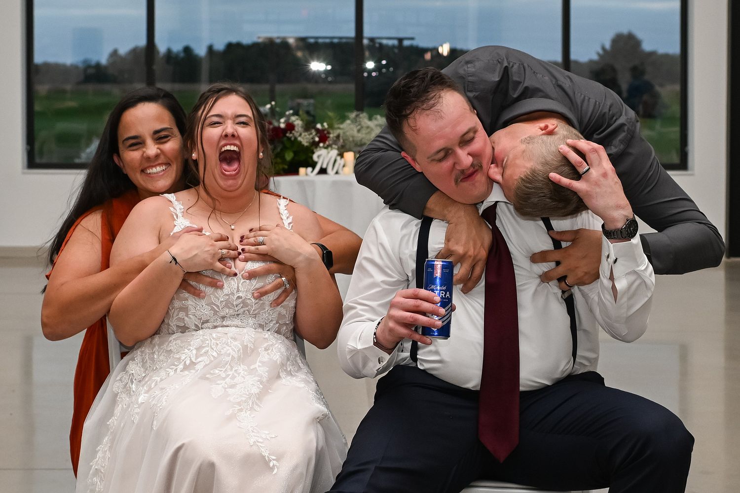 Joyful wedding party celebrating with drinks, bride laughing while groom gets kissed on cheek by friend.