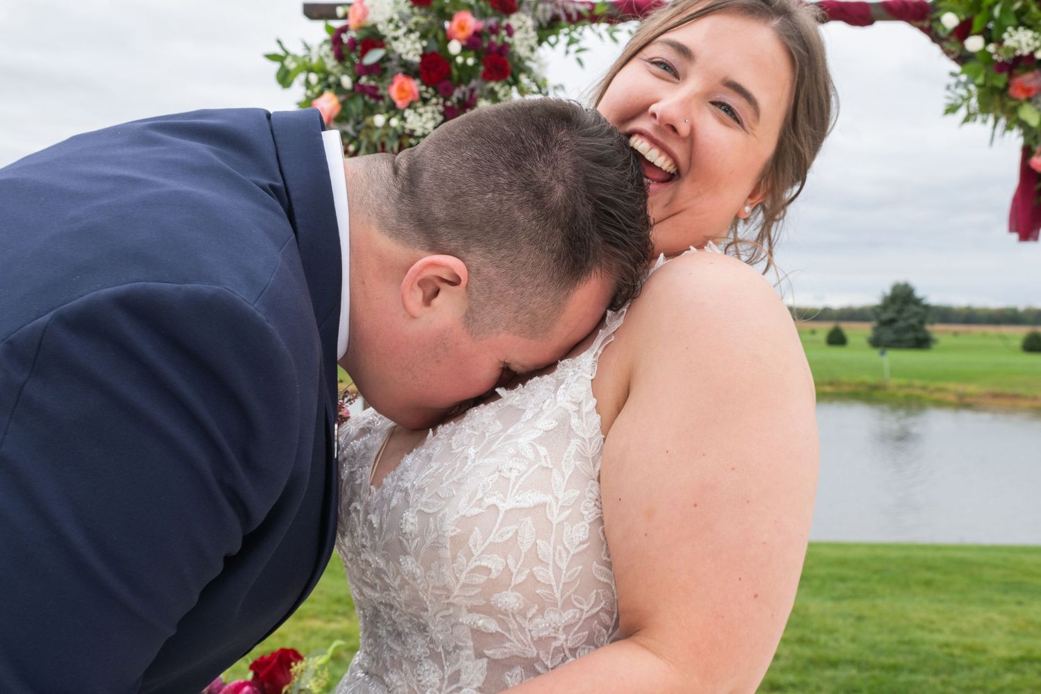 Bride laughing joyfully as groom kisses her shoulder during outdoor wedding ceremony by a pond.