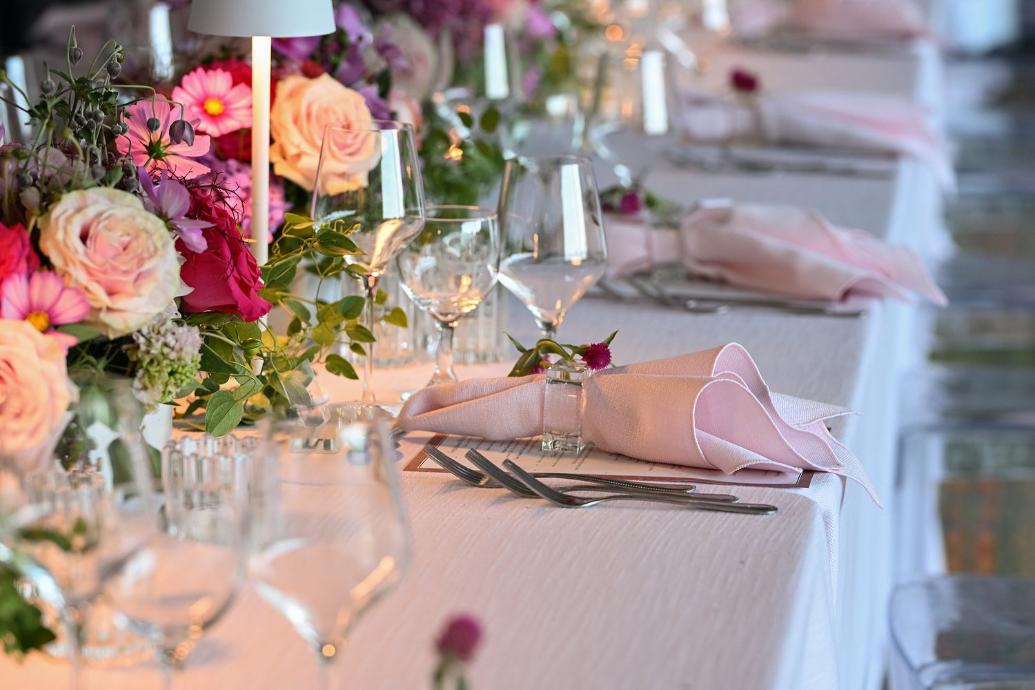 Elegant wedding table setting with peach roses, pink napkins, wine glasses and glowing candles in romantic lighting.