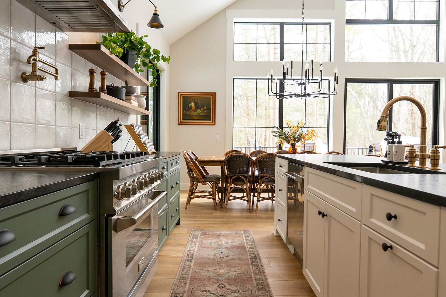A farmhouse kitchen with green range, open shelves, white cabinets, black countertops, and a dining area with chandelier.