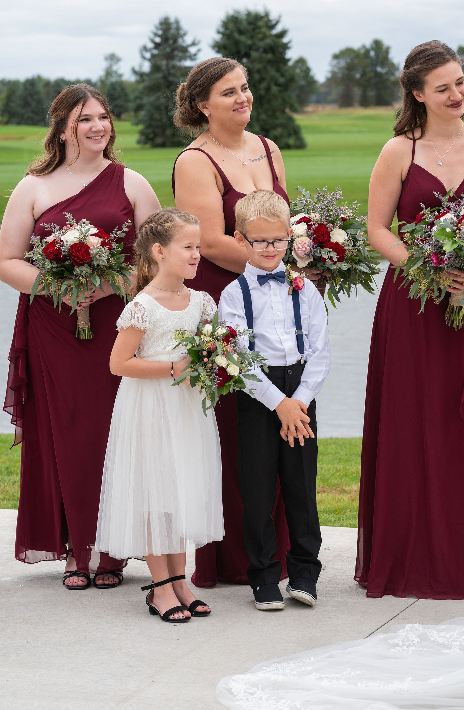 Bridesmaids in burgundy gowns hold red floral bouquets beside flower girl in white dress and ring bearer in suspenders.