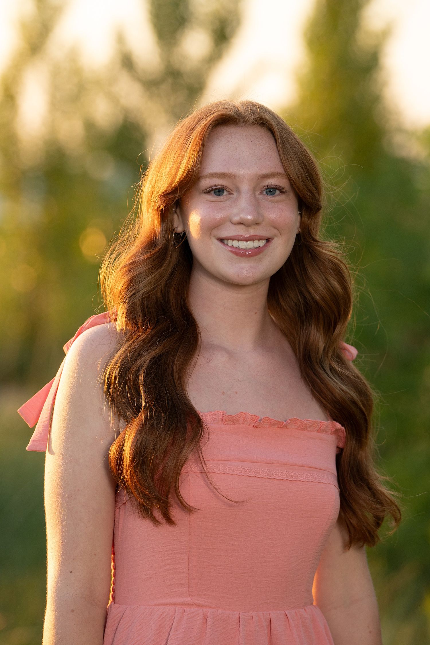 Smiling young woman with long red hair wearing a pink dress outdoors in golden hour light.