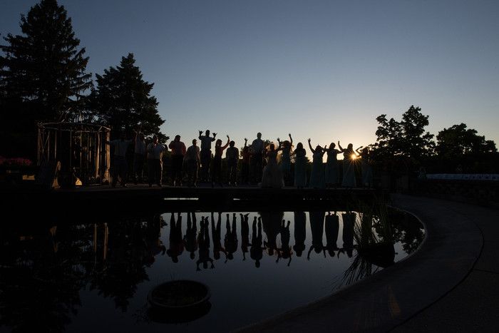 Silhouettes of wedding party reflected in water at sunset with trees in background.