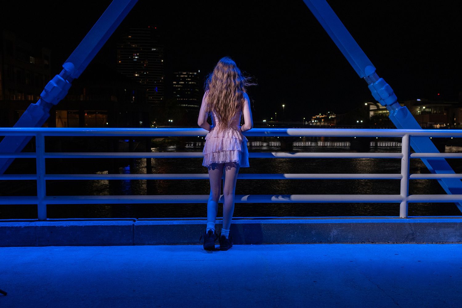 Someone in a dress stands at the railing of a bridge illuminated in blue light.