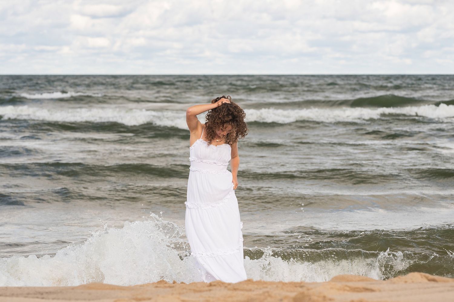 Woman in white dress plays in ocean waves on cloudy beach day.