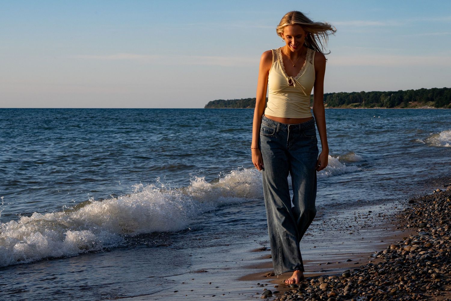 Person in casual attire walking along a rocky beach shoreline with waves crashing at sunset.