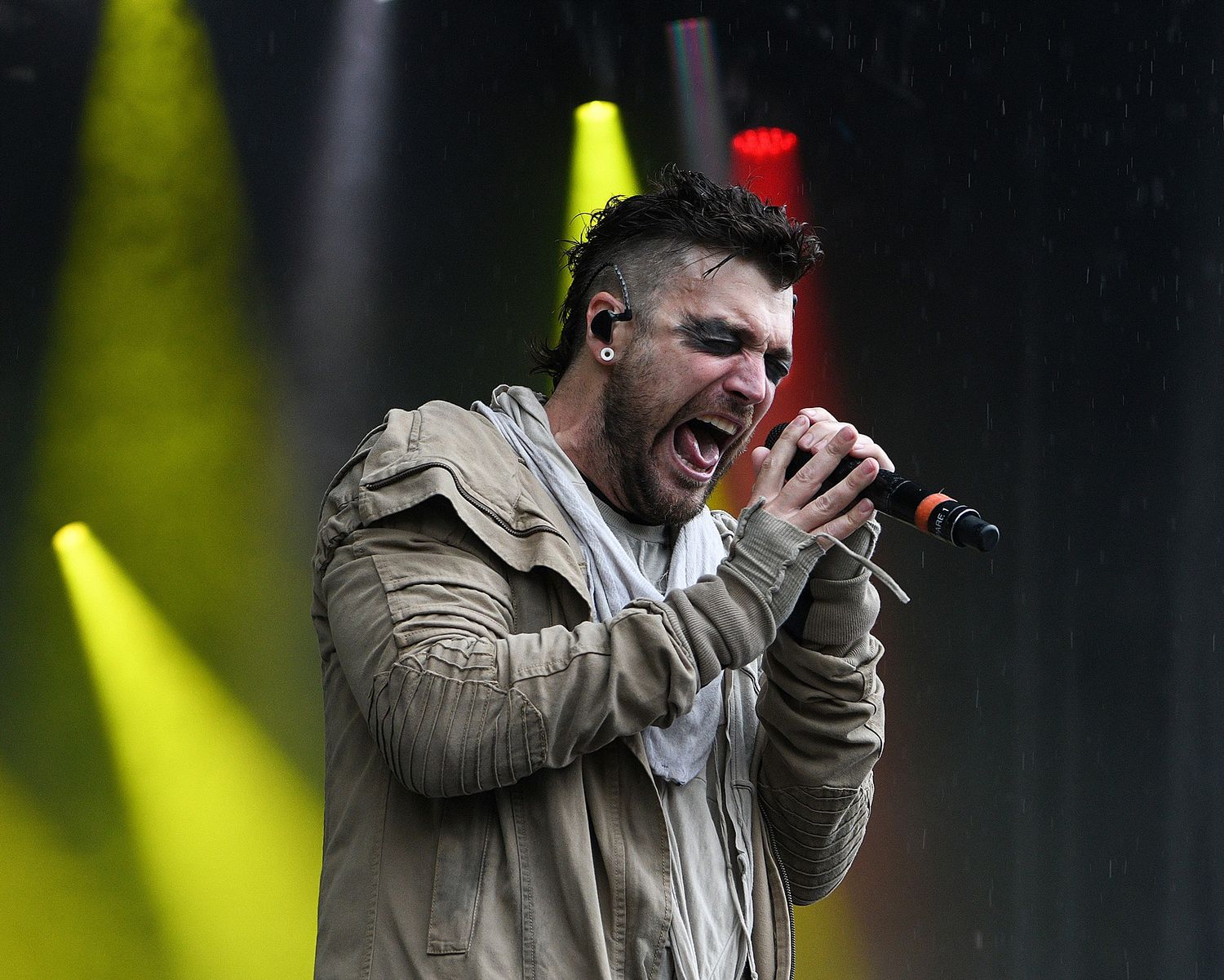 Singer in beige jacket performing intensely with microphone under colorful stage lights.