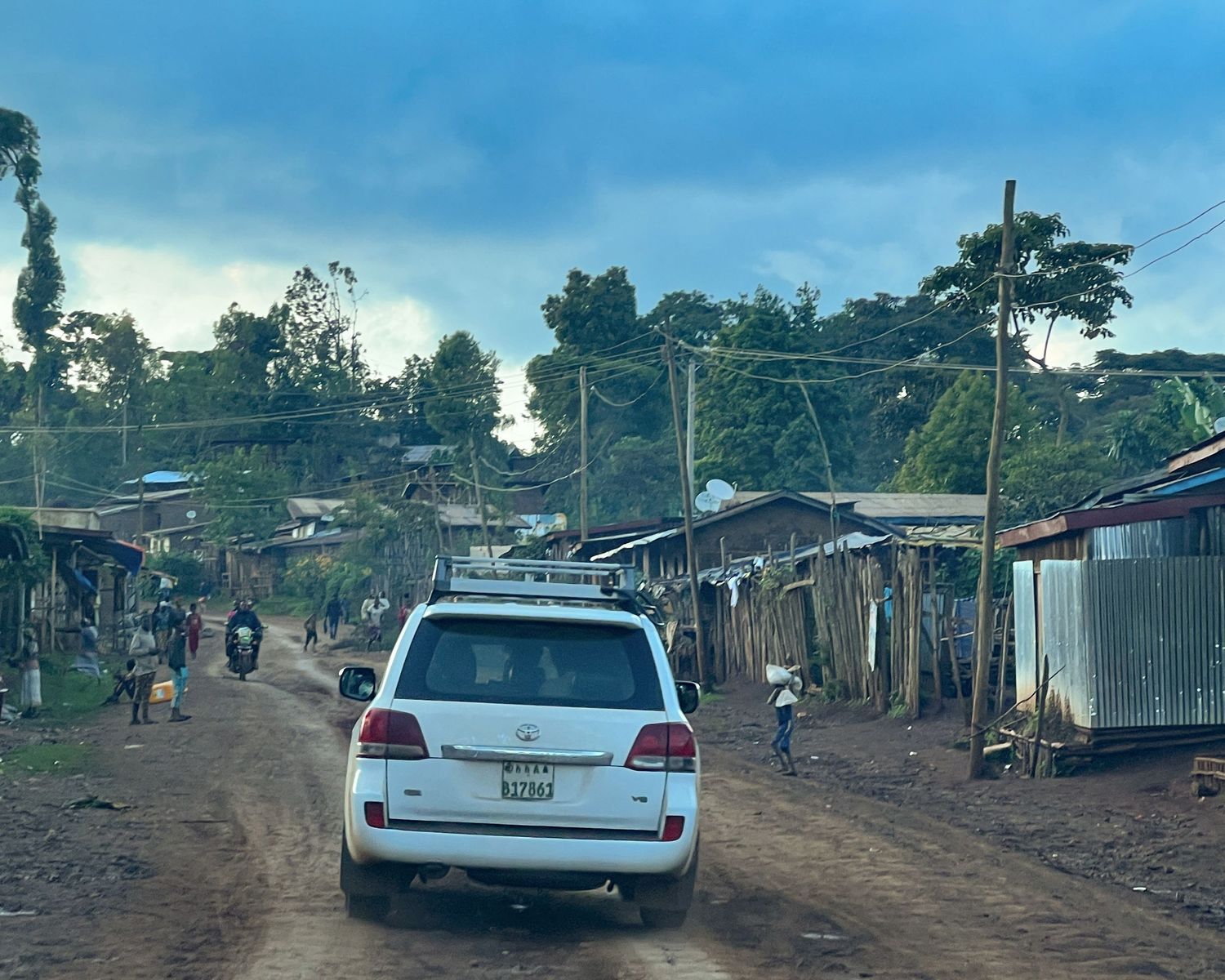 Rural village street with white cars parked along basic housing structures.