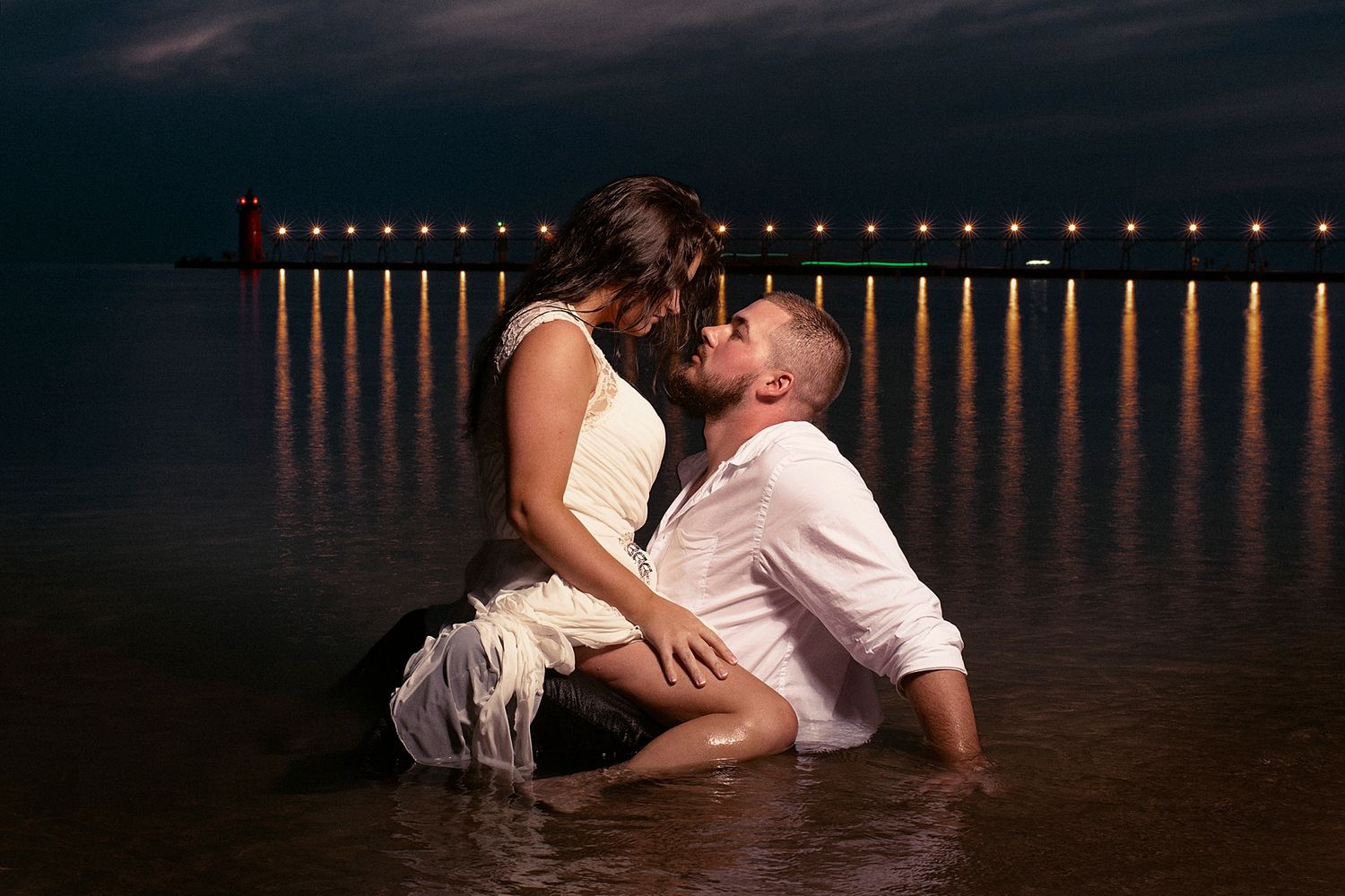 Couple in formal attire posing in water at night with illuminated pier in background.