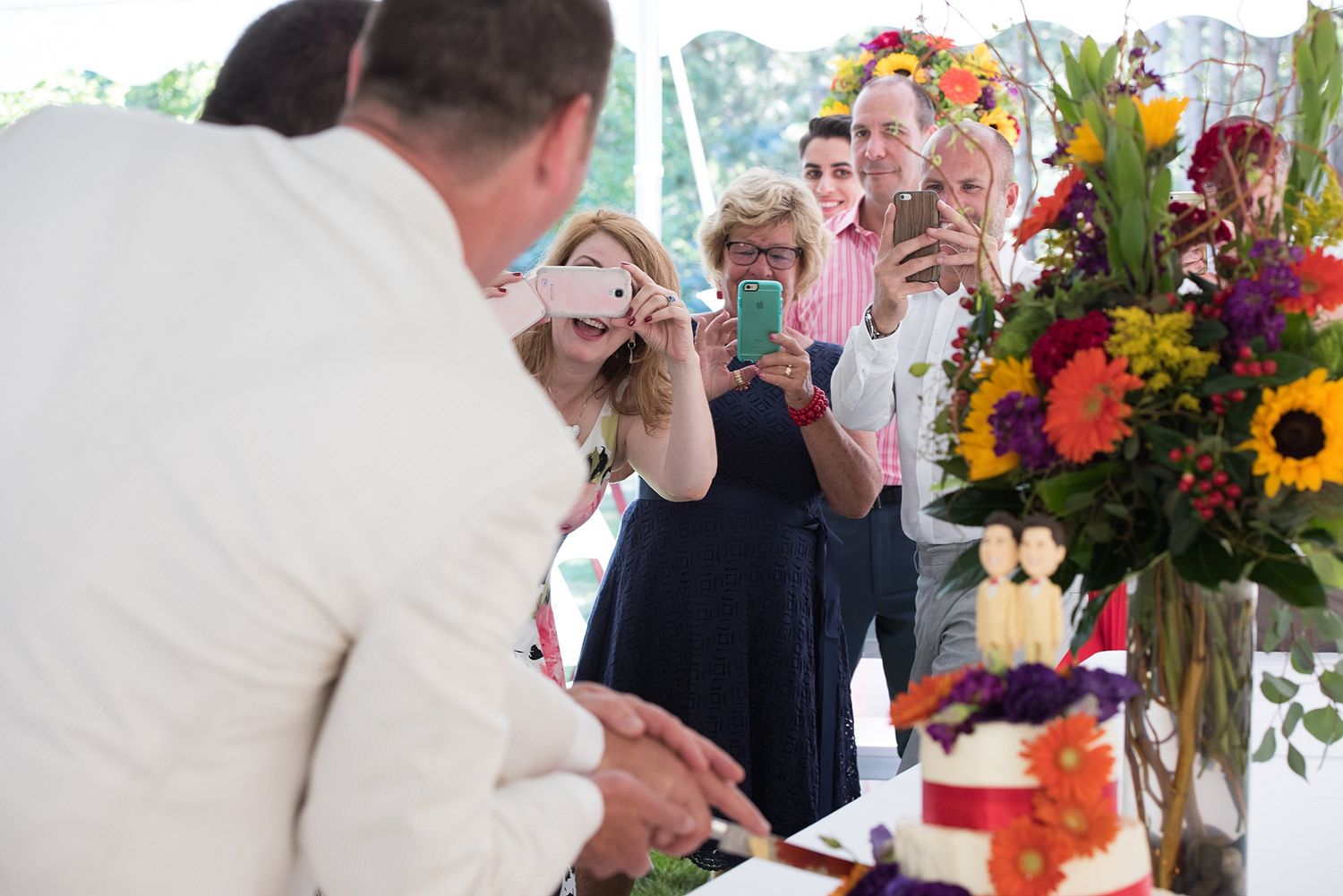 Guests photograph a wedding cake cutting ceremony under a tent with colorful floral arrangements nearby.