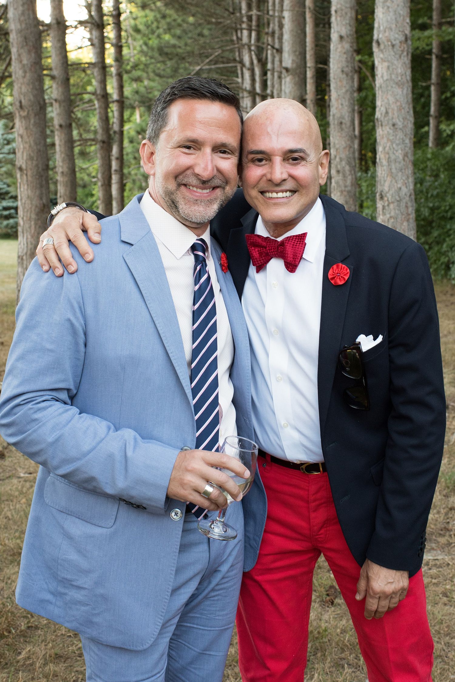 Two well-dressed men in blazers smiling together outdoors, one in red pants and bow tie, holding a wine glass.
